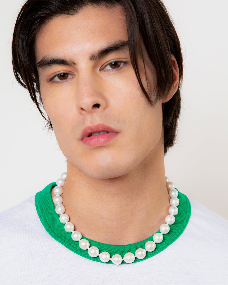 A man wearing a strand of large white pearls at the base of his neck, over a white T-shirt with a green collar