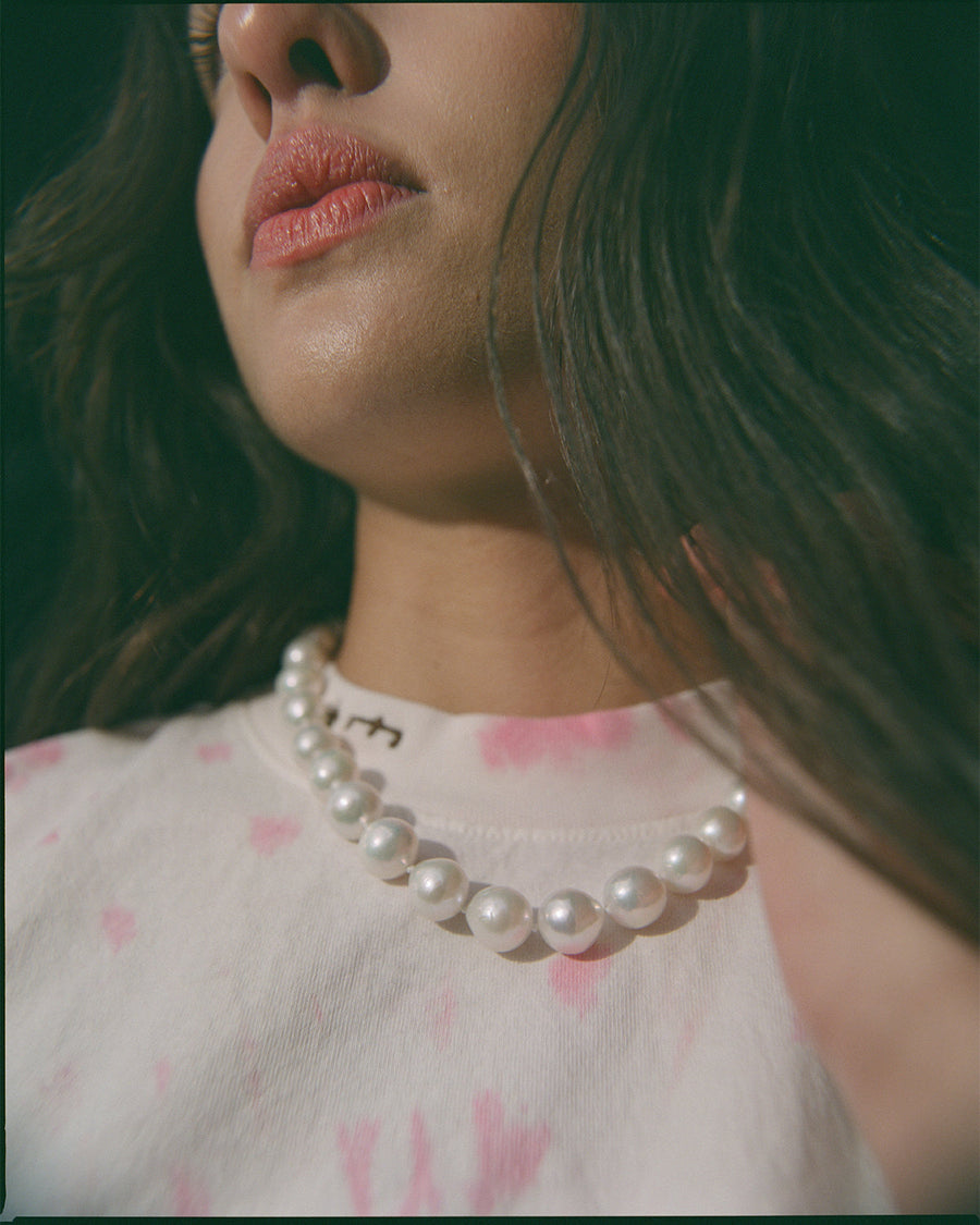 A woman with her face turned up to the sun, wearing a strand of large white pearls at the neck of a pink and white T-shirt