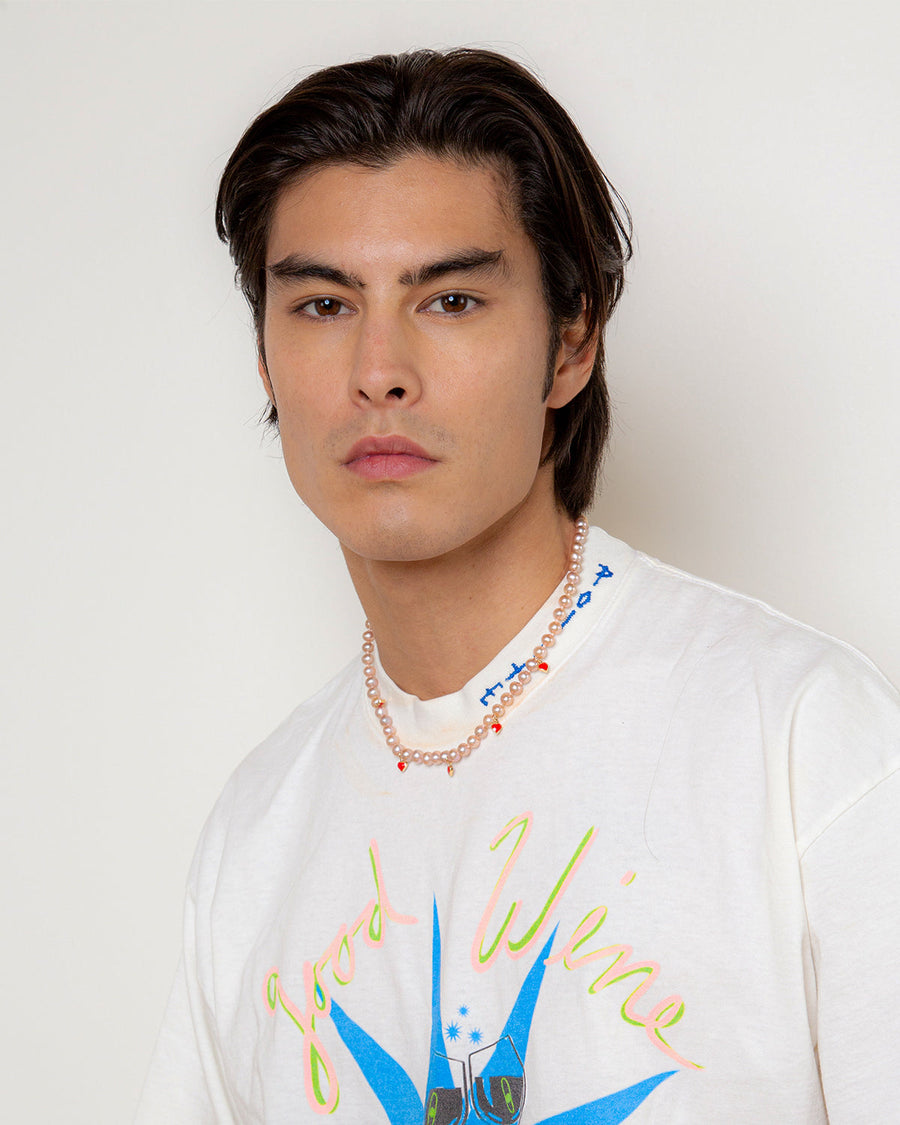 A man in a white graphic T-shirt wearing a strand of pink pearls with small red heart charms at the base of his neck